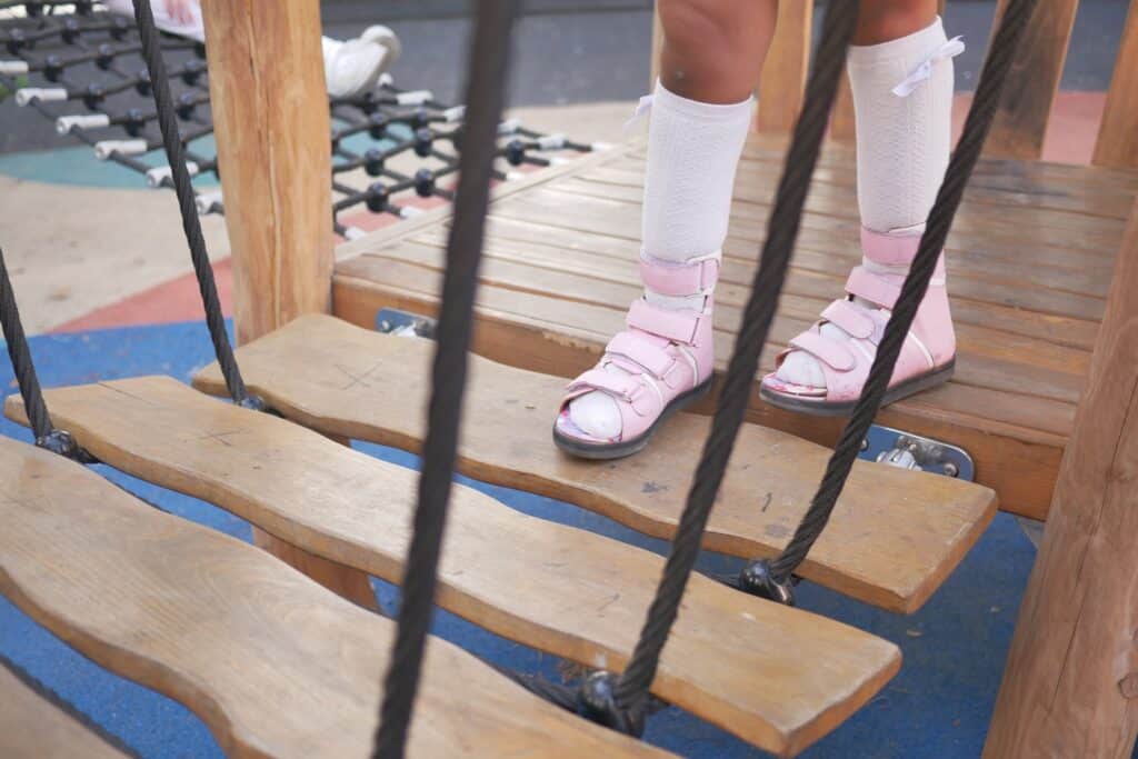 A little girl in orthosis shoes crosses a wooden bridge Child cerebral palsy disability.